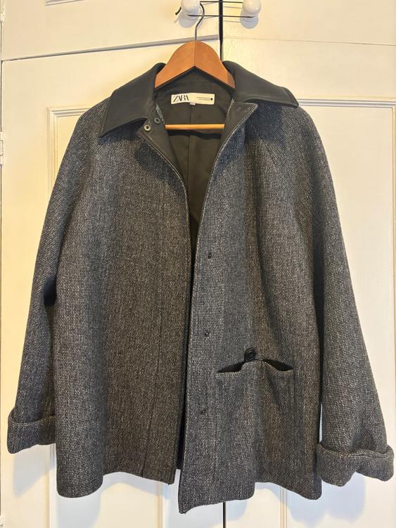 Textured grey coat hanging on a wooden hanger, with a dark collar and a visible label that reads "ZARA". White door background.