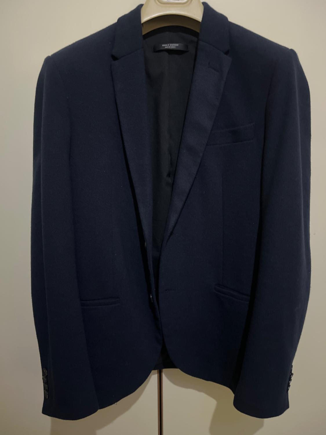Navy blue jacket hanging on a beige hanger, with classic lapels and front pockets. Plain white background.