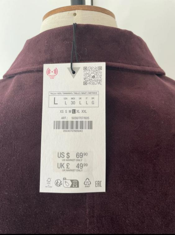 Tag hanging from a burgundy coat on a mannequin. Shows sizes and prices: US $69.90, UK £49.99. QR code and barcode visible.