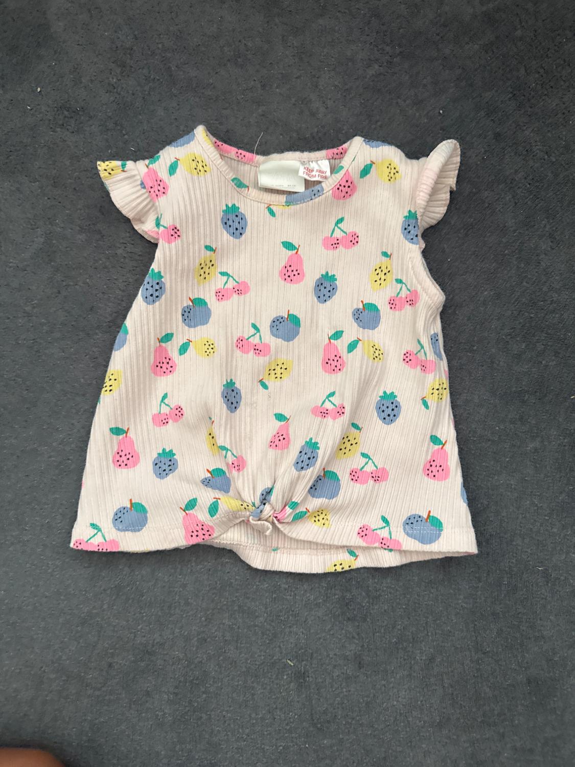 Pink ribbed fruit-print T-shirt with ruffled sleeves and a knotted hem. Visible text: KEEP AWAY FROM FIRE.