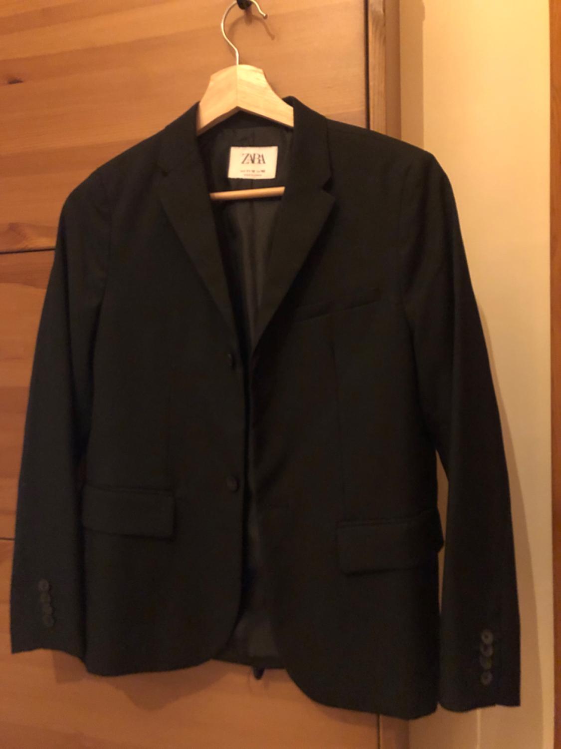 Black jacket hanging on a wooden hanger, with visible label that says "ZARA". Light wood background.