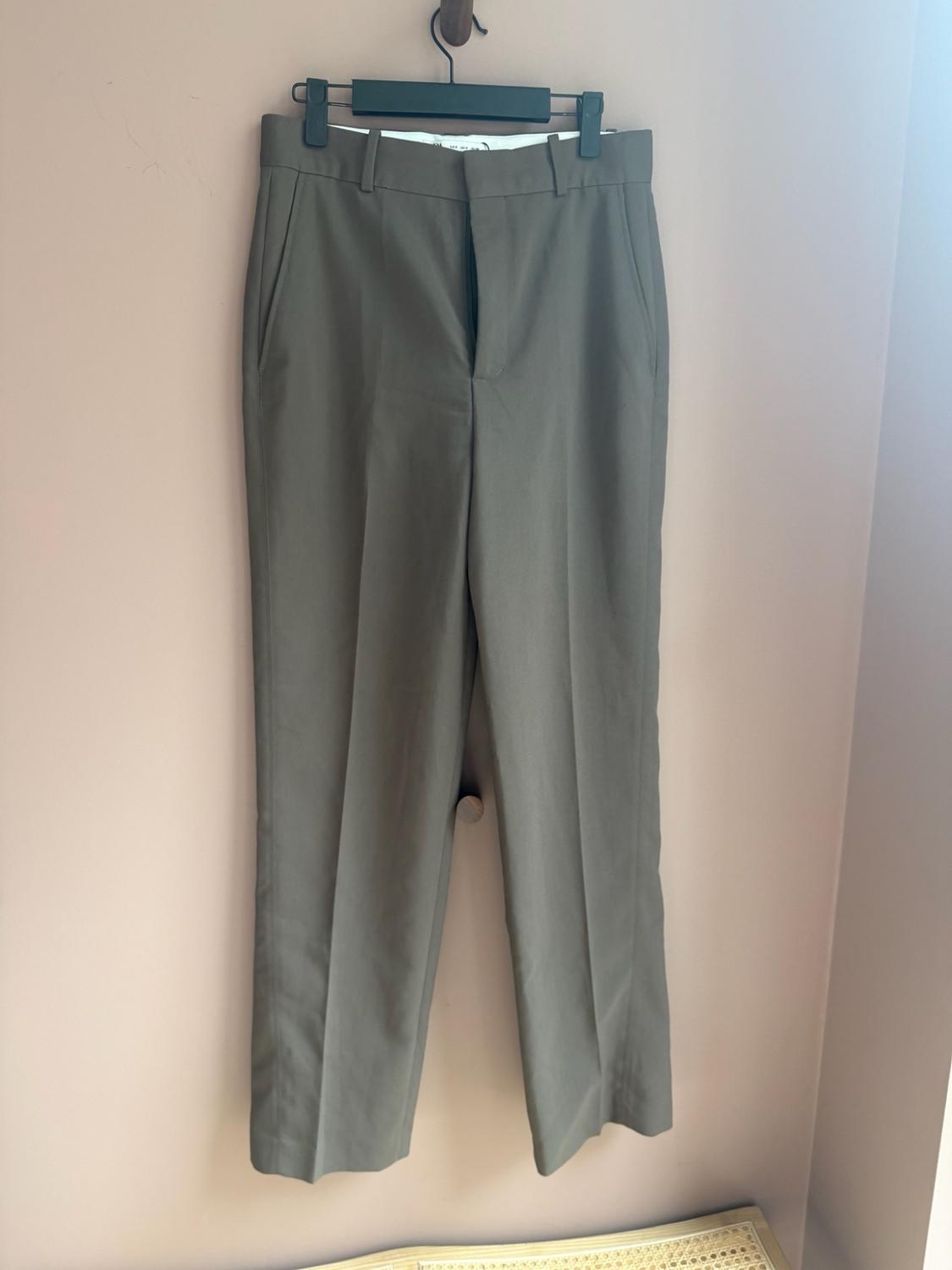 Khaki green dress trousers with a front crease, hanging on a black hanger against a pale pink wall.