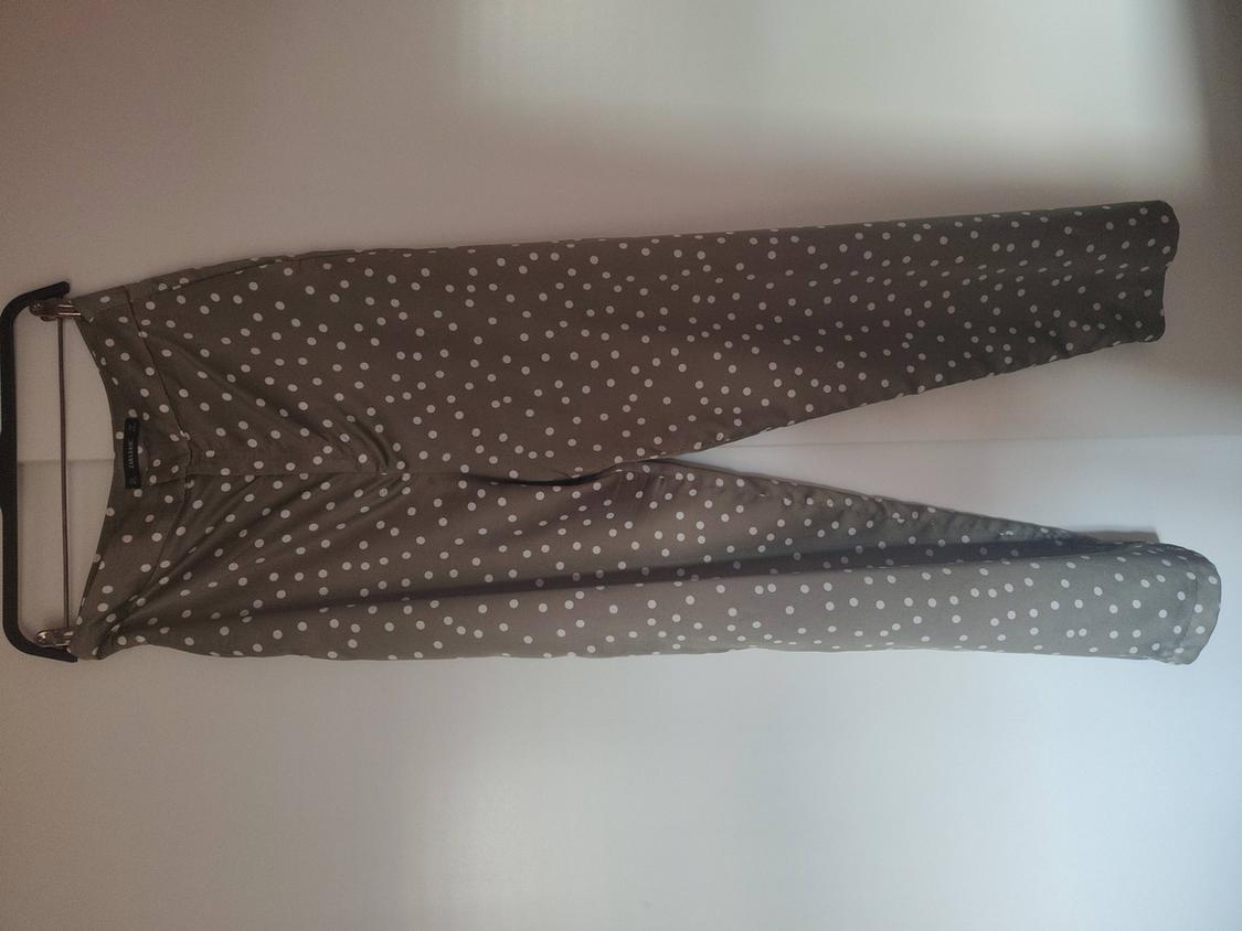 Olive green trousers with a white polka dot print, hanging from a black hanger with clips against a white background.
