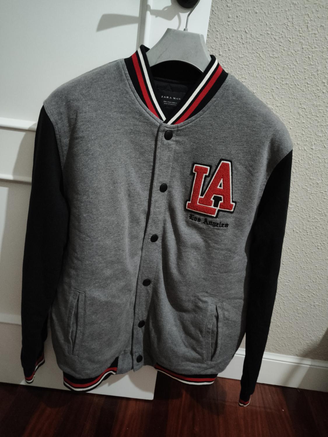 Grey varsity jacket with black sleeves, front buttons and red "LA Los Angeles" embroidery, hanging on a hanger in front of a beige wall.