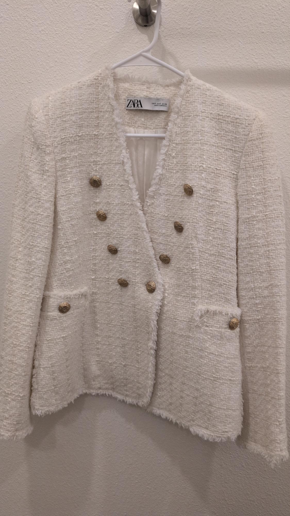 White textured fabric jacket with gold buttons and two front pockets, hanging on a white hanger. Visible label: ZARA.