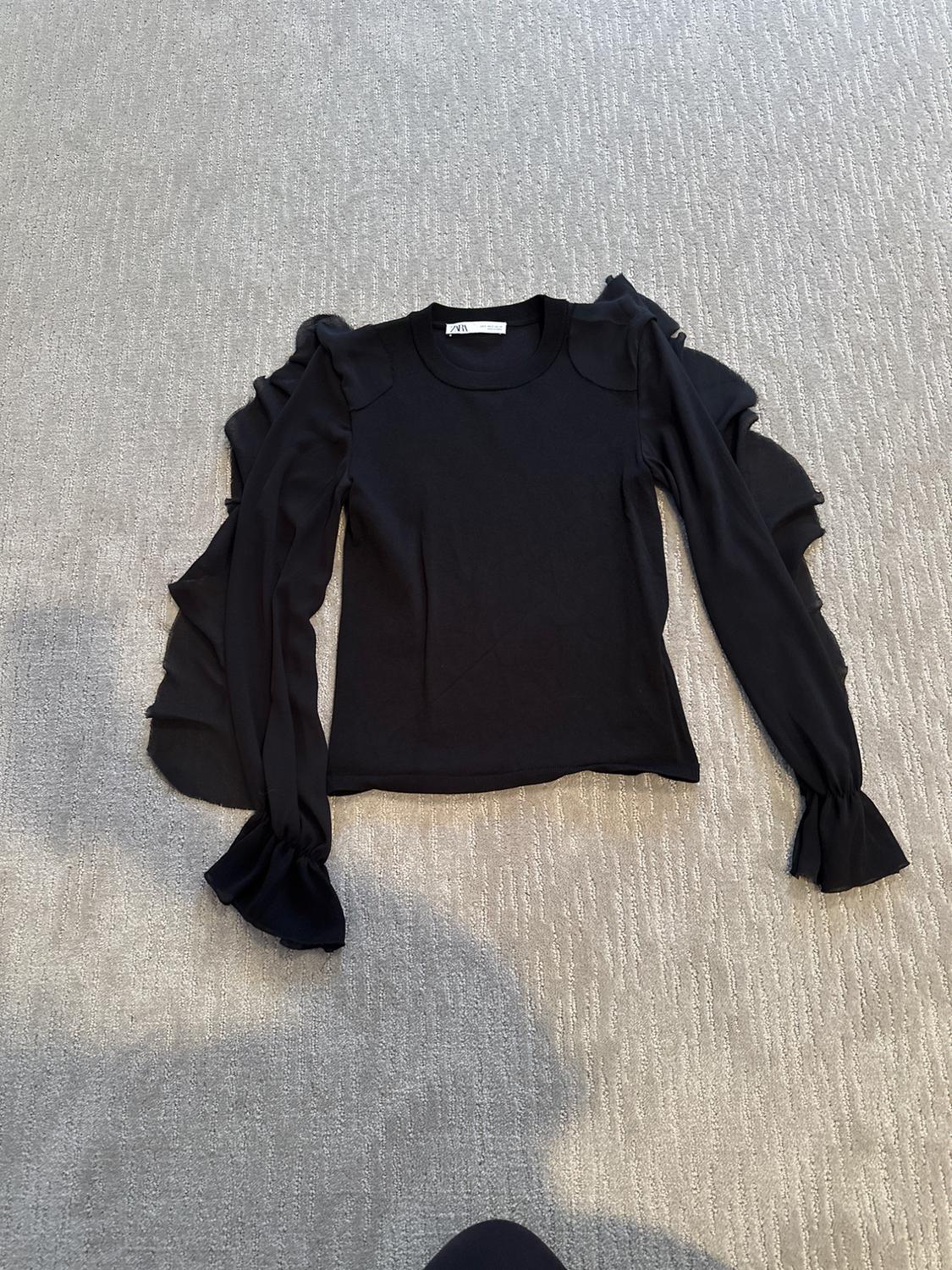 Black long-sleeved jersey with ruffles on the sleeves, on a gray rug. Visible tag on the collar.