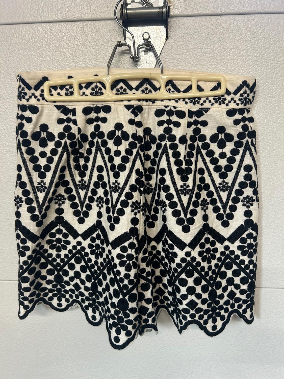 White shorts with black zigzag embroidery and floral motifs, hanging on a hanger. Featuring a scalloped hem.