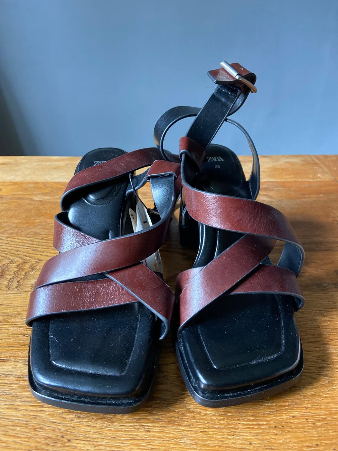 Heeled sandals with a square black sole and brown crossover straps on a wooden table. Visible text: ZARA 36.