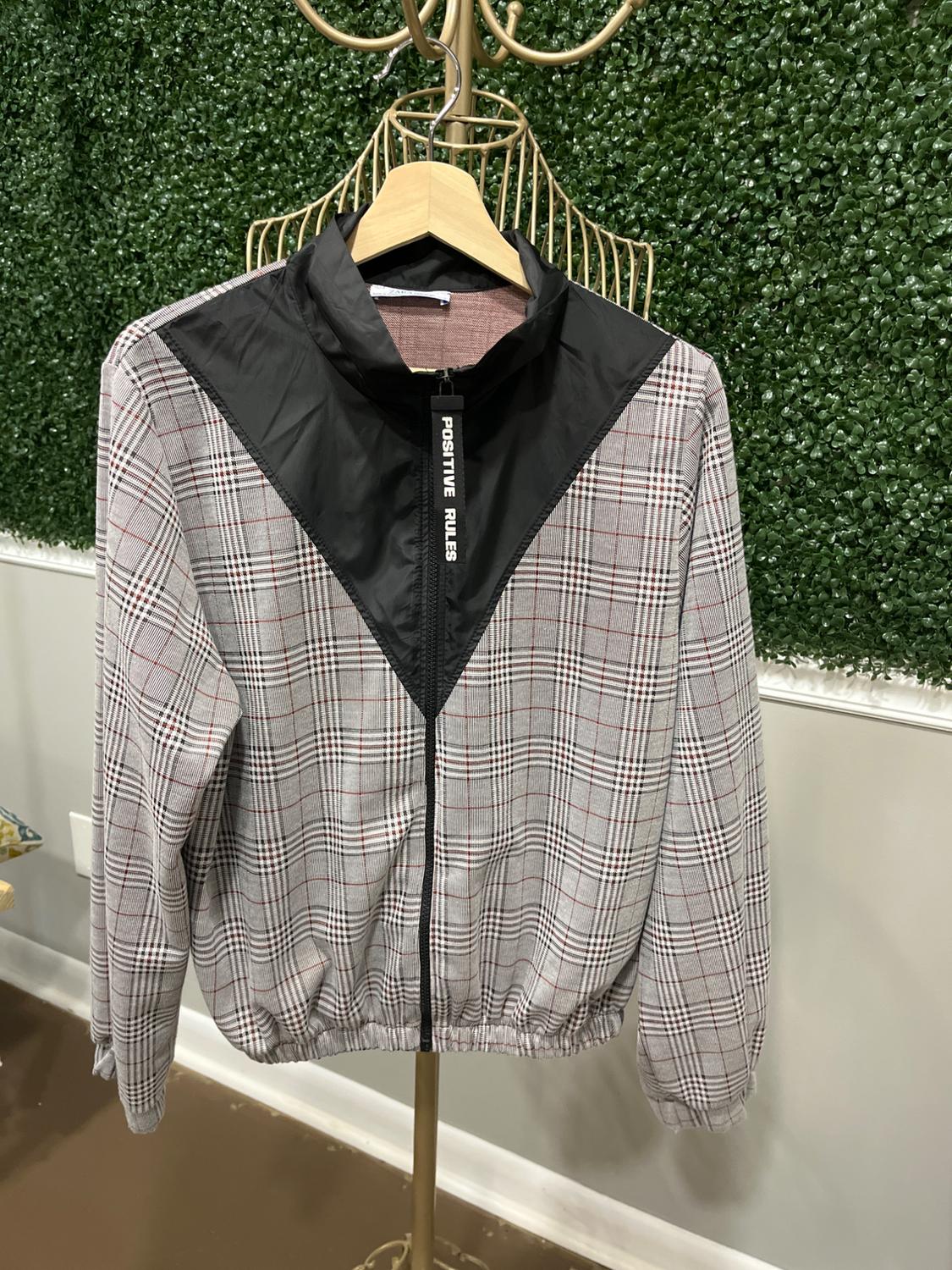Plaid jacket with a black chest panel, "POSITIVE RULES" text on the zipper, hanging on a gold clothes rack.