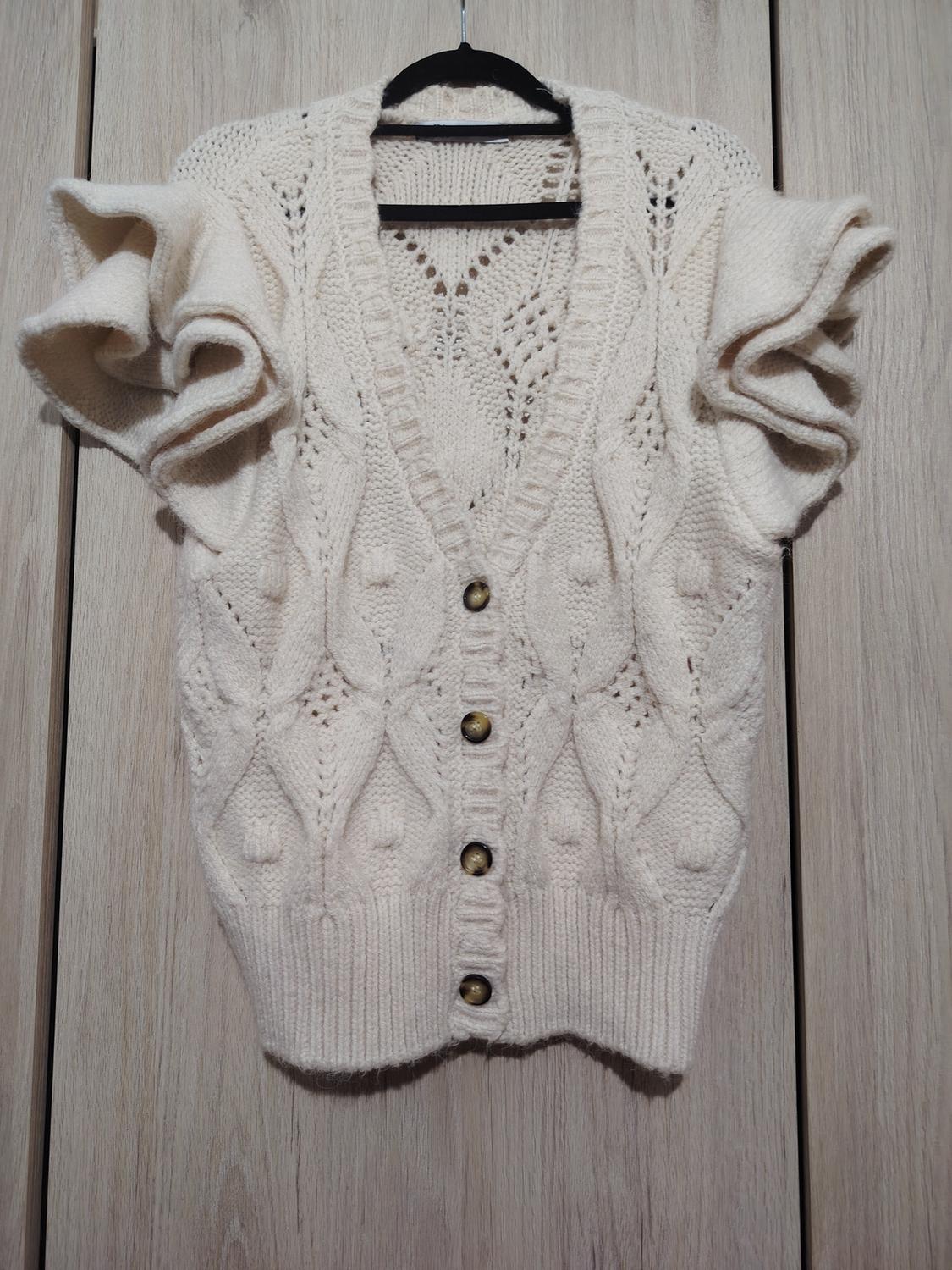 White knitted cardigan with gold buttons and puff sleeves, hanging on a hanger against a light wooden background.