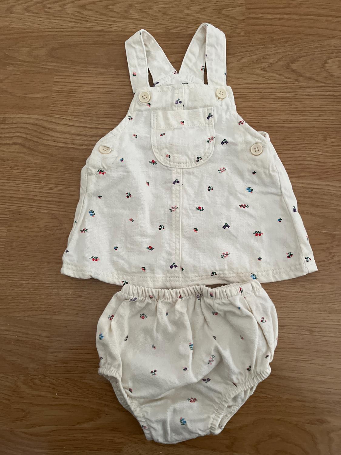 Conjunto infantil blanco con estampado de flores pequeñas, compuesto por un peto con tirantes y botones, y un pantalón corto. Fondo de madera.