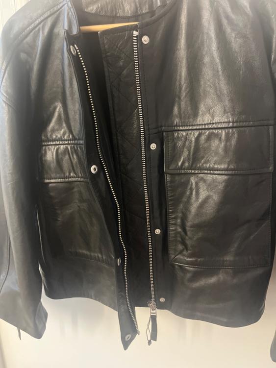 Black leather jacket hanging on a hanger, with an open zipper and front pockets. White background.