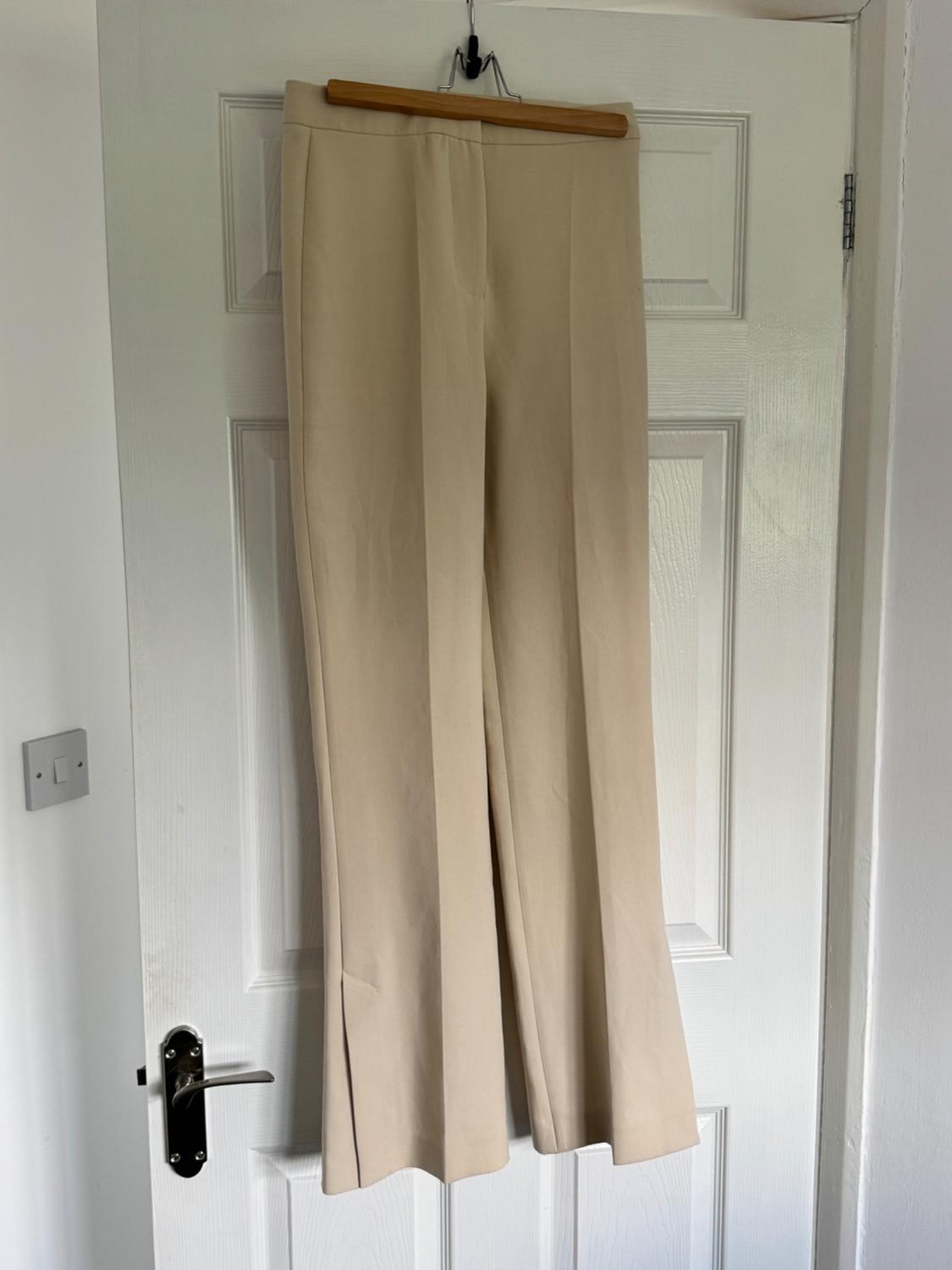 Beige trousers hanging on a wooden coat hanger over a white door. The door has a silver-coloured door handle.