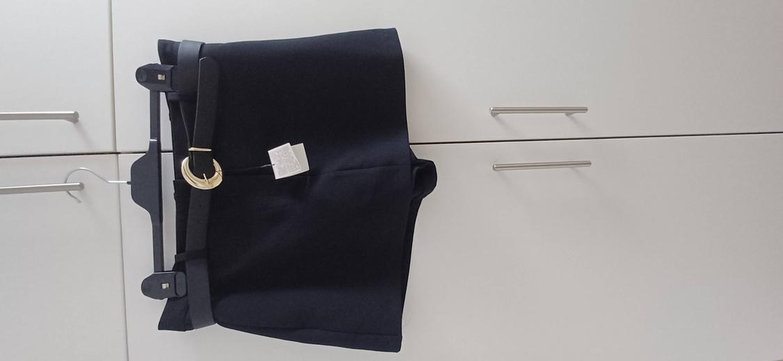 Black shorts with a belt and circular gold buckle, hanging on a hanger in front of a white wardrobe.