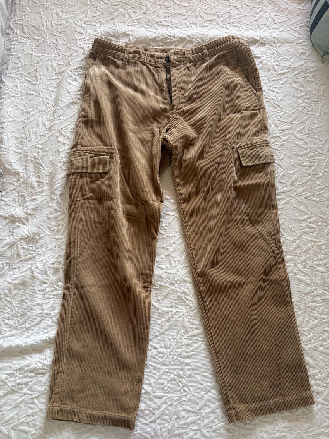 Light brown corduroy cargo trousers with side pockets, on a white bedspread with floral texture. Casual and relaxed style.