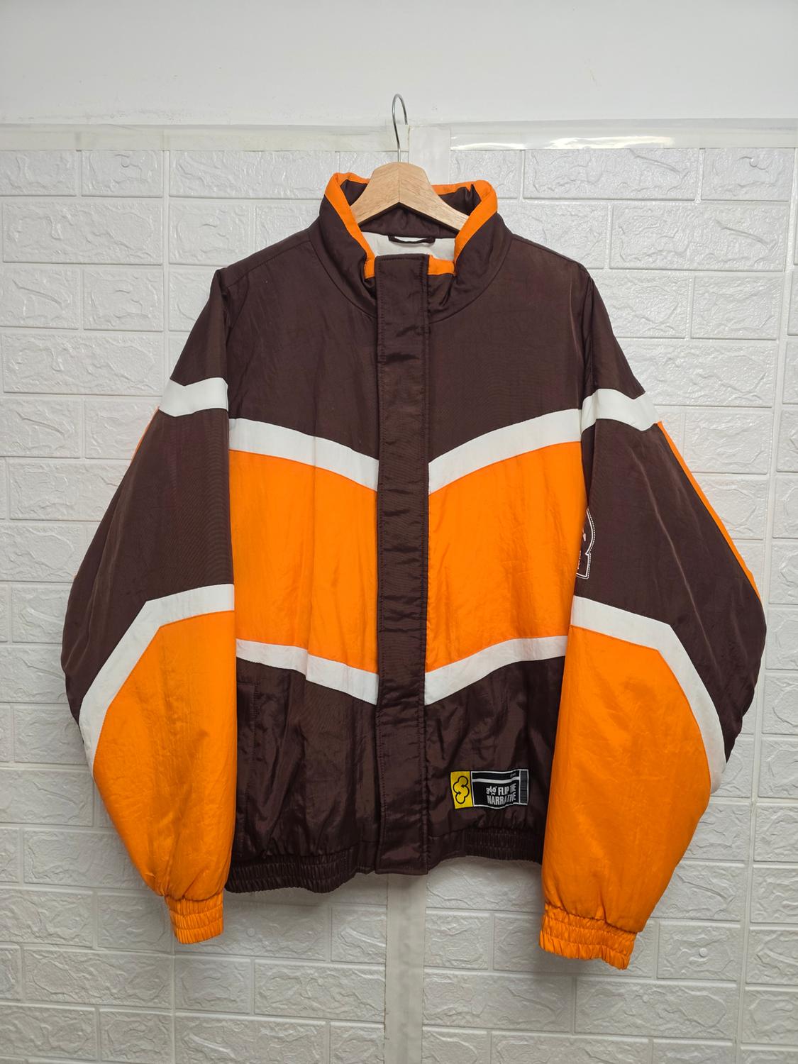 Jacket hanging on a coat hanger, with a colour block design in brown, orange and white. White wall background with a brick texture.