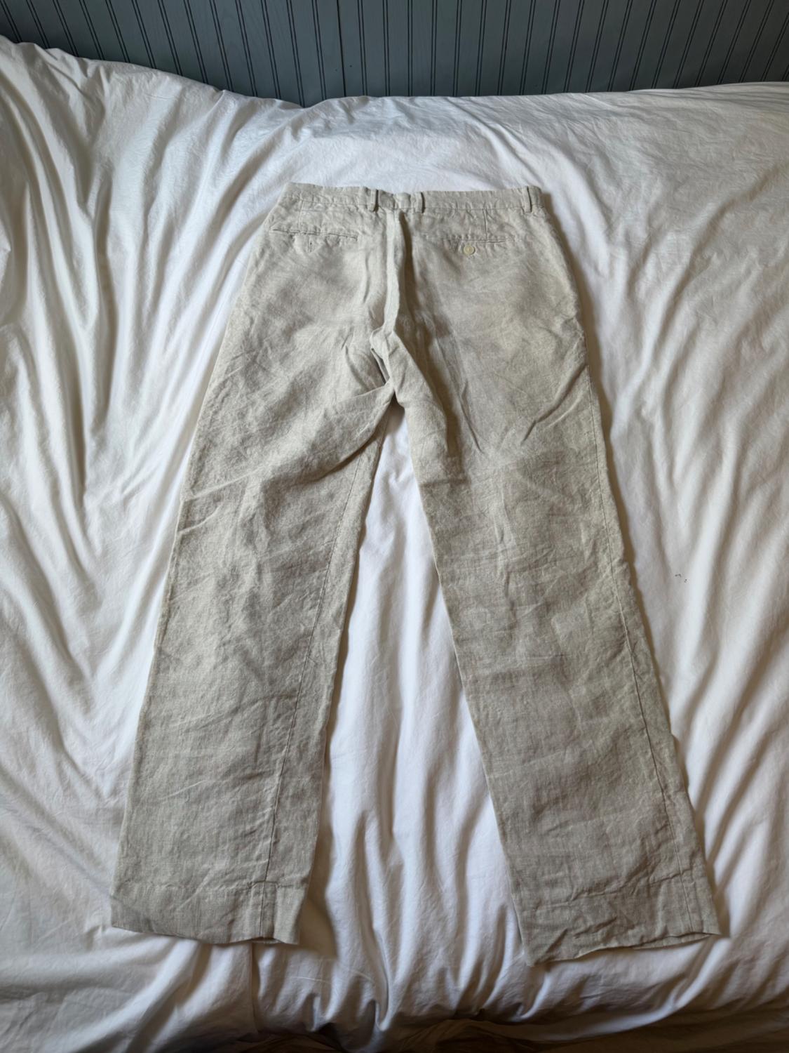 Beige pants seen from behind, spread out on a bed with wrinkled white sheets. Relaxed atmosphere with a gray headboard in the background.