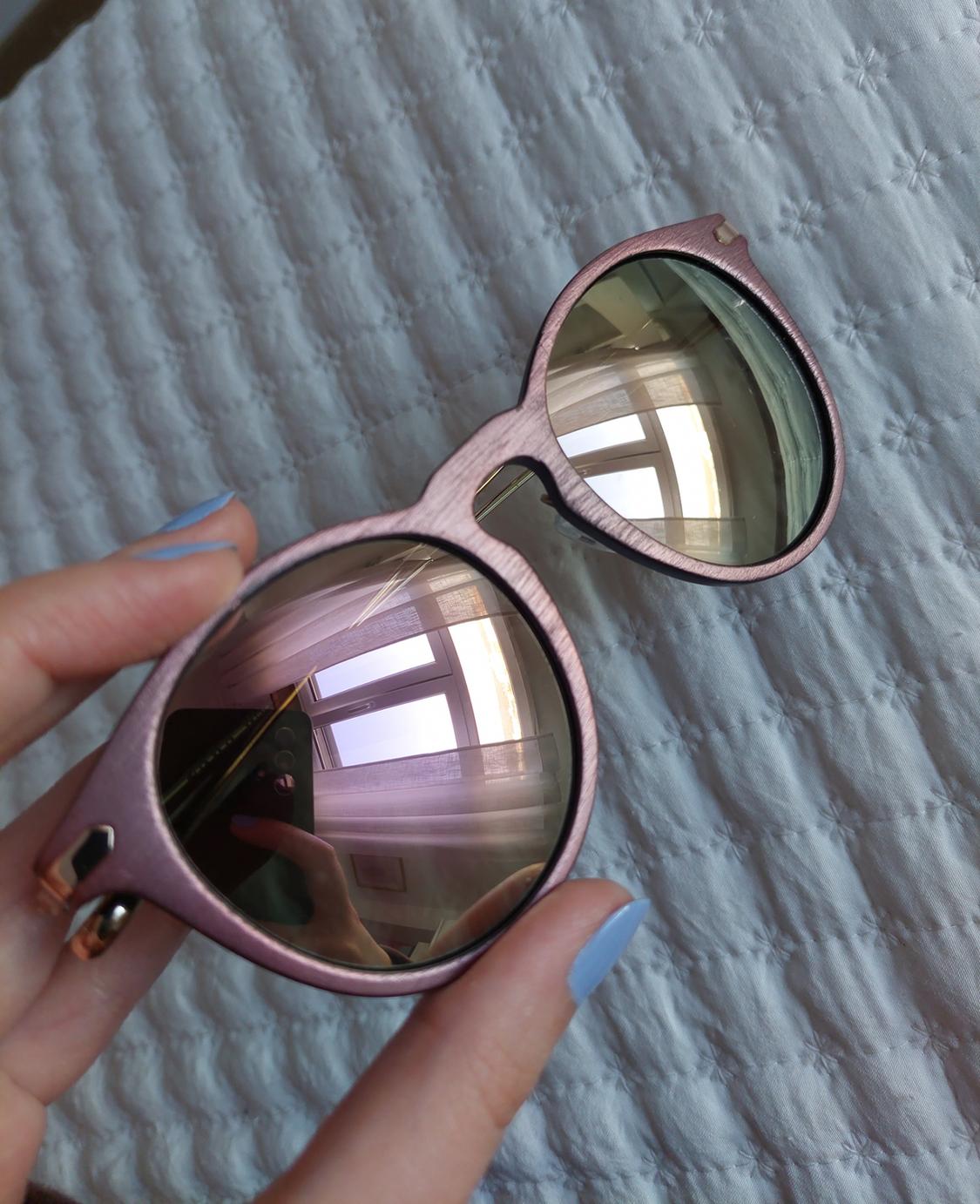 A hand with blue nails holding metallic pink-rimmed sunglasses with mirrored lenses over a quilted white fabric.