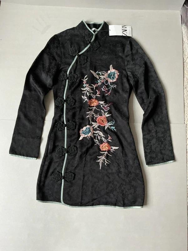Black dress with floral embroidery in pink and green tones, decorative buttons, and visible tag. Light grey background.