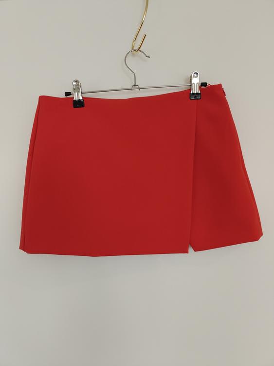 Red short skirt hanging on a metal hanger with black clips, on a white background.