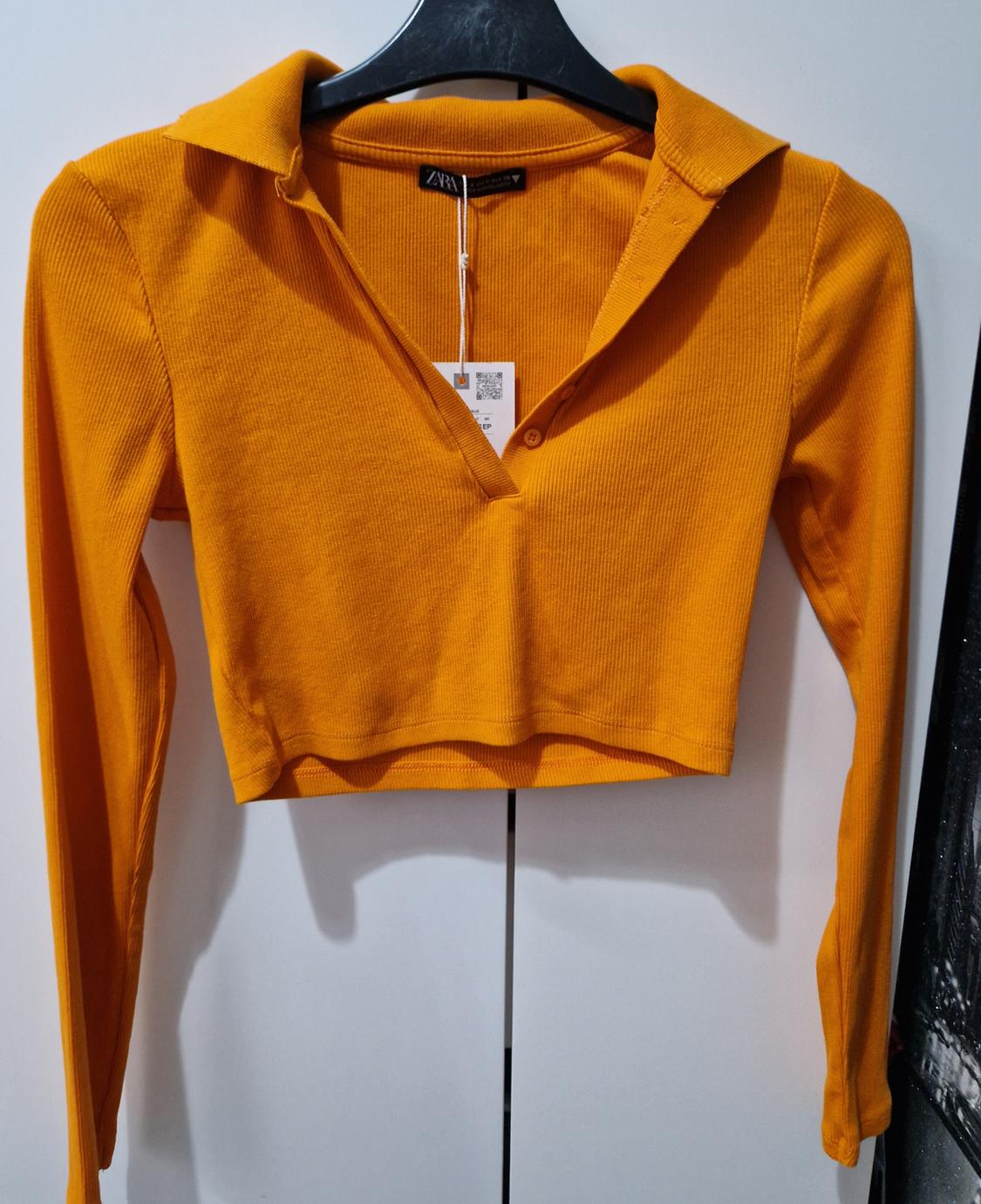 Orange ribbed long-sleeved crop top with a polo neck and V-neckline. Visible ZARA label. Hanging on a hanger against a white background.