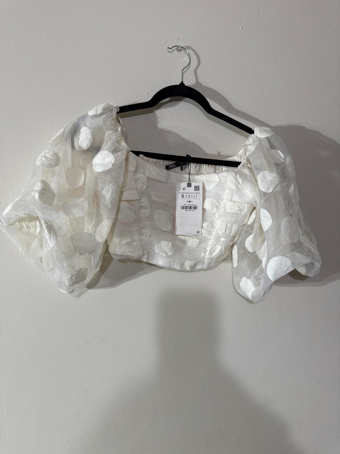 White blouse with puff sleeves and a circle print, hanging on a black hanger against a white wall. Label visible.