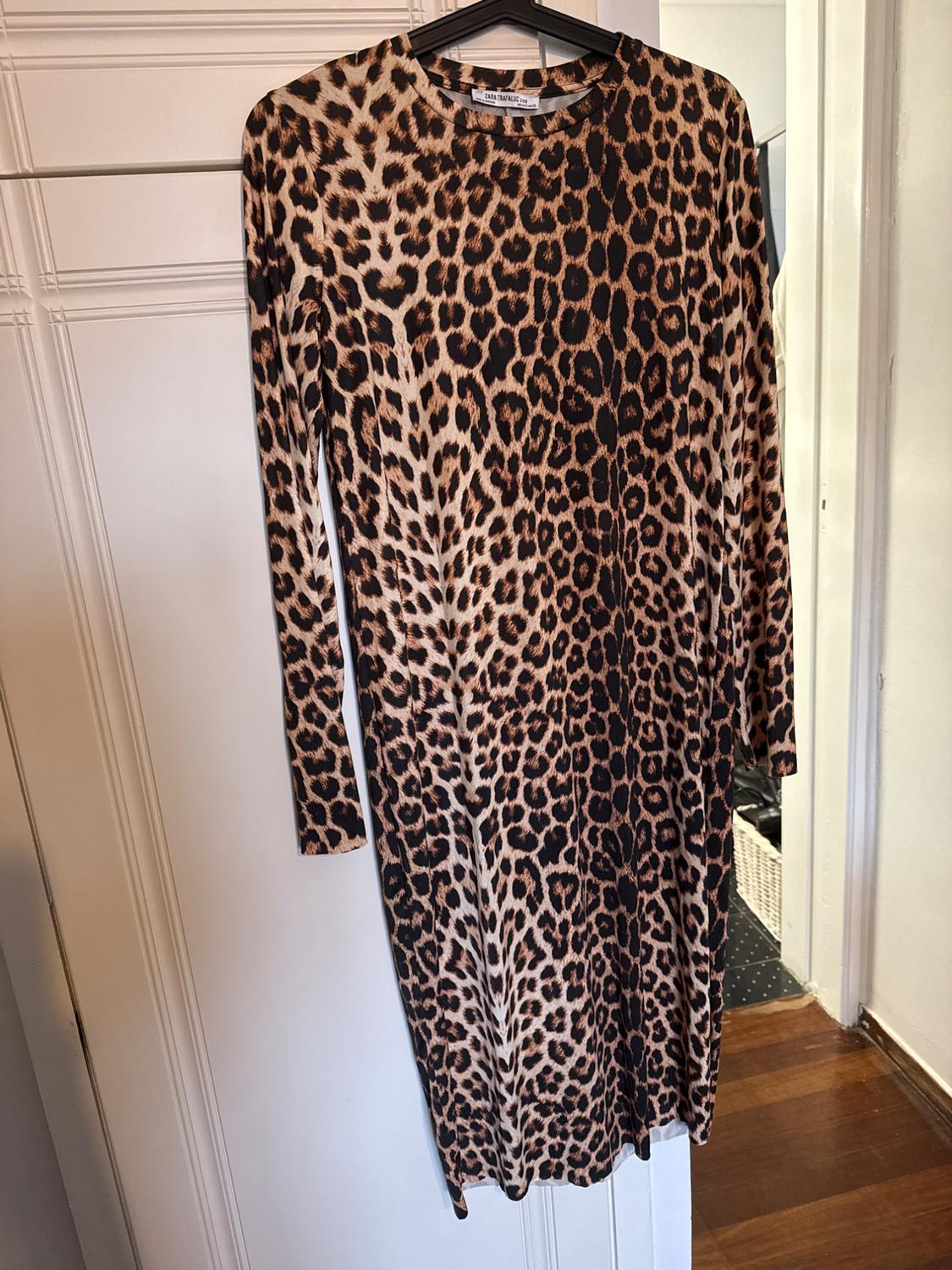 Long-sleeved leopard print dress hanging on a hanger, against a white wall and wooden floor.
