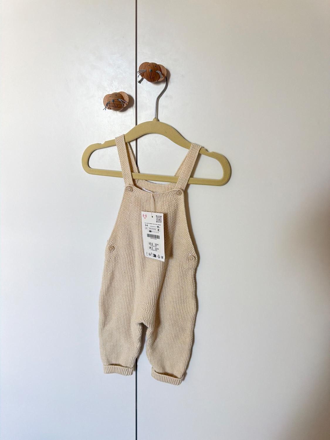 Beige knitted babygrow hanging on a yellow hanger over a white door. Label visible in the centre of the babygrow.