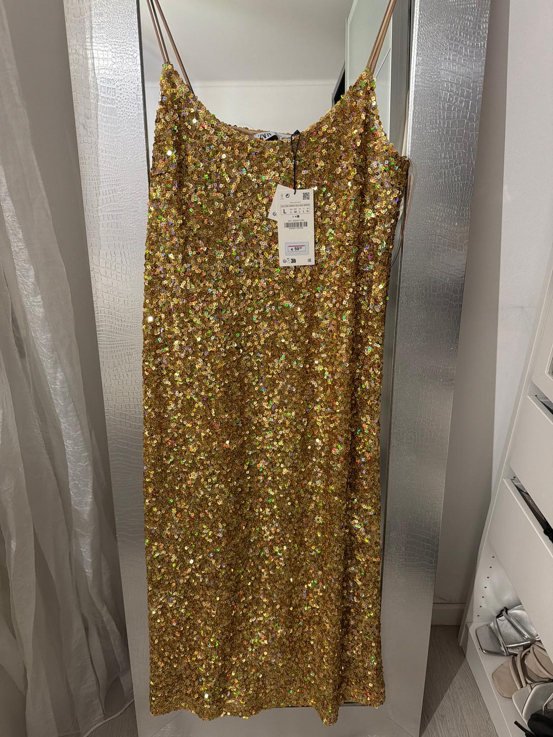 Gold sequinned dress hanging on a mirror, with visible tags. Room background with curtains and shelves.