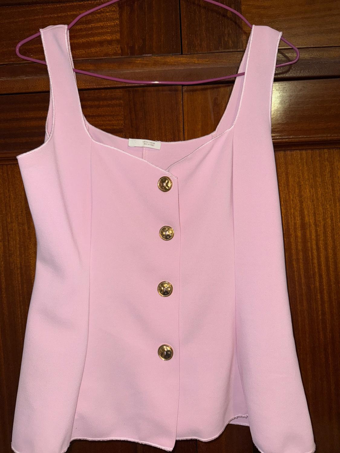 Light pink top with wide straps and five gold buttons at the front, hanging on a pink hanger against a wooden background.