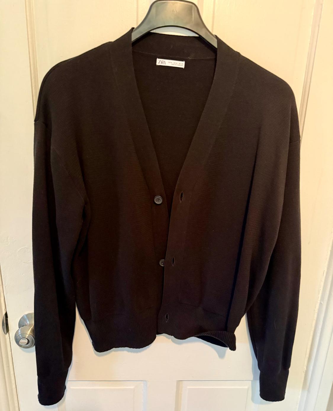 Black knitted cardigan hanging on a hanger over a white door. Label visible on the neck.