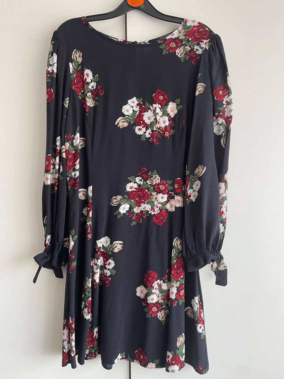 Black dress with a floral print in shades of red, white and green, long sleeves with tie cuffs, hanging on a hanger.