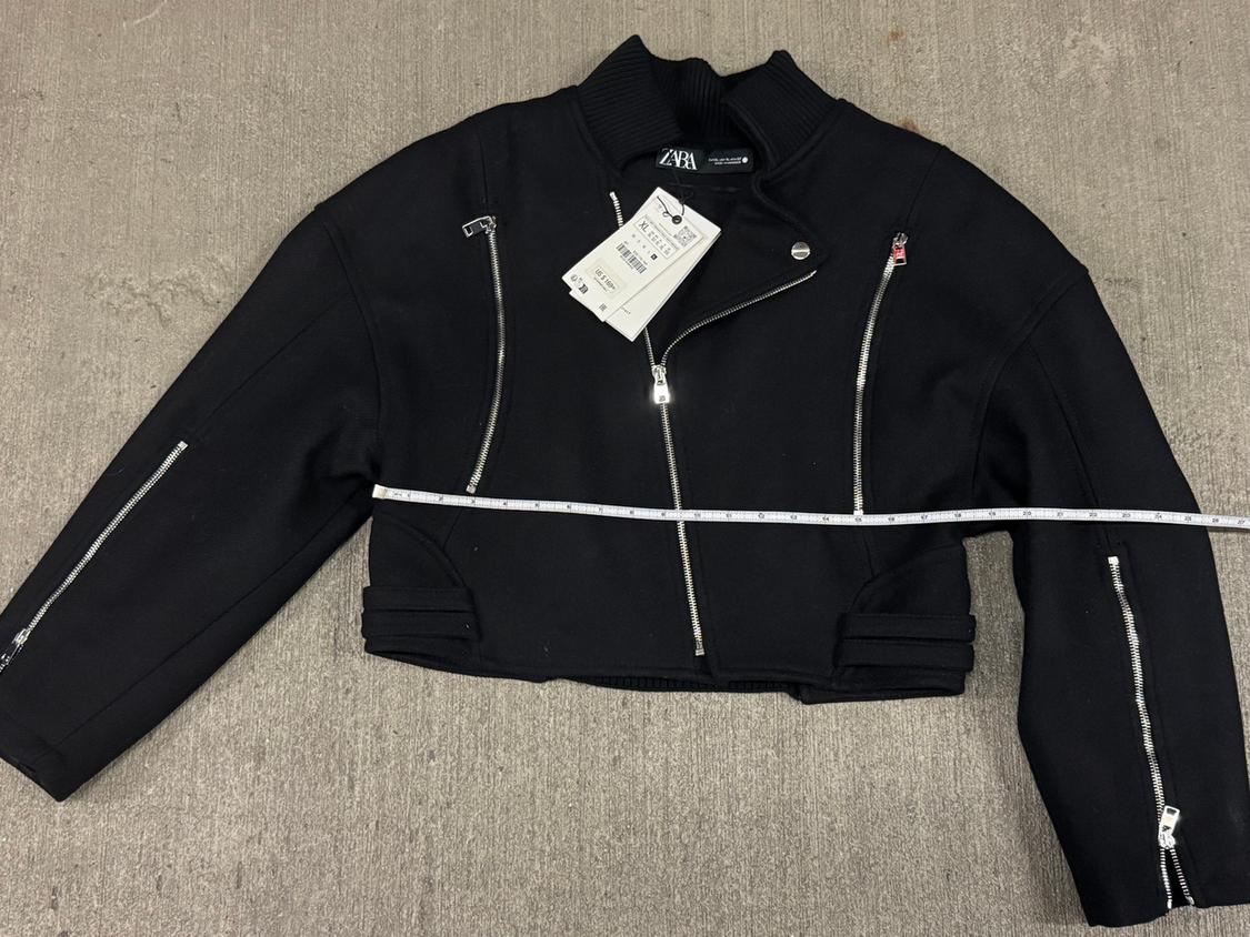 Short black jacket with silver zippers and a visible tag on a gray background. A measuring tape measures the width.