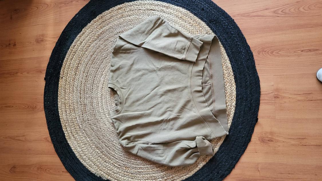 Beige long-sleeved sweatshirt on a circular jute and black rug, on a light wooden floor.