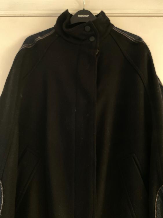 Black coat hanging on a Topshop hanger, with a high collar and buttons at the top, visible stitching on the shoulders.