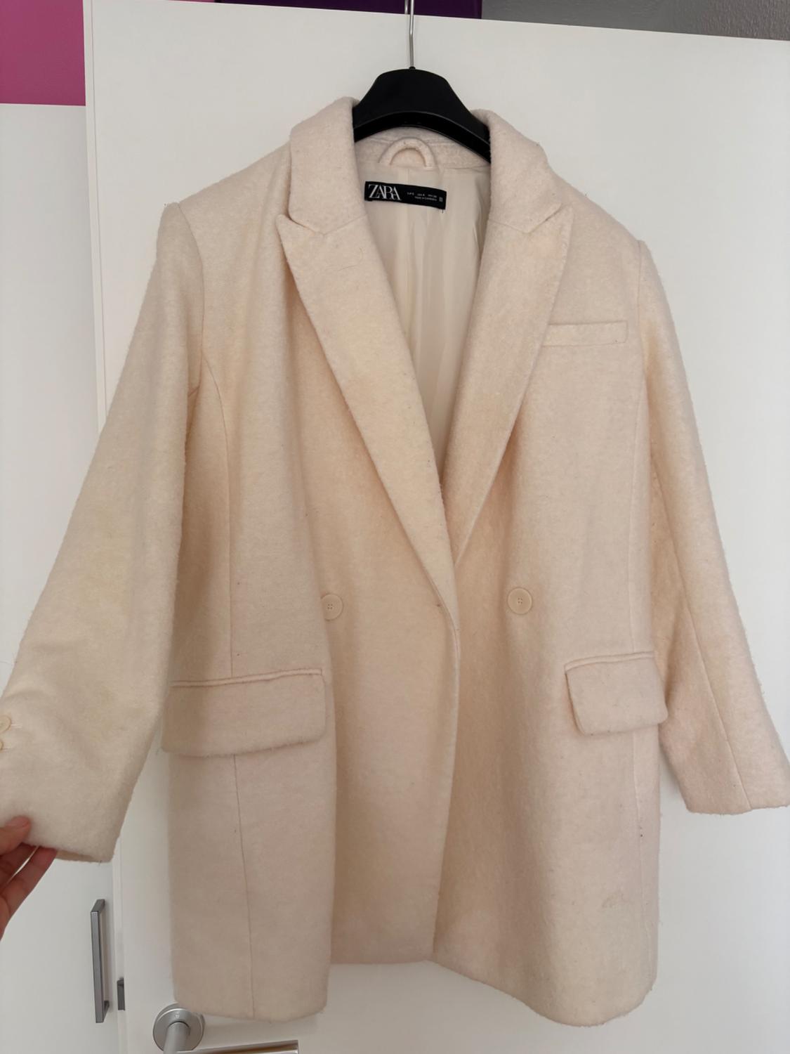 Beige jacket hanging on a black hanger, with lapels and front pockets. White and pink background. Visible label with "ZAPA" text.