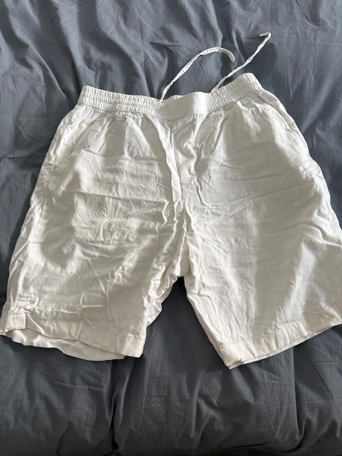 White crinkled shorts with an elasticated drawstring waist, laid on a bed with grey sheets.