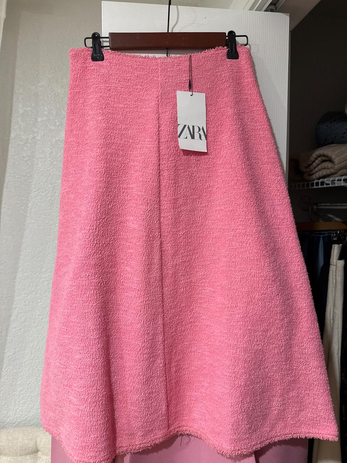 Pink textured skirt hanging on a hanger with a visible "ZARA" tag, white door background, and shelves with clothes.