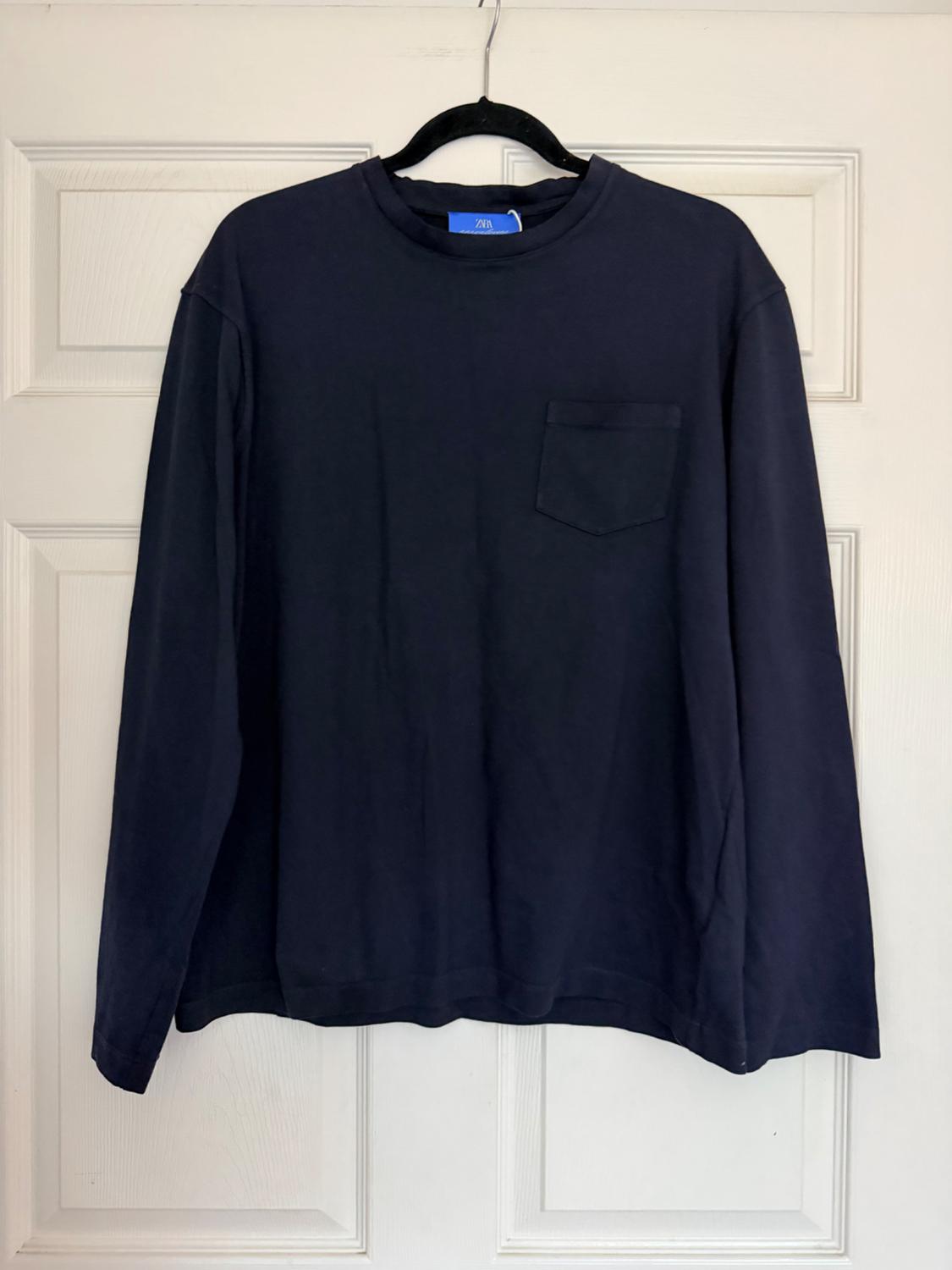 Navy blue long-sleeved shirt with a chest pocket, hanging on a hanger over a white door. ZARA label at the neck.