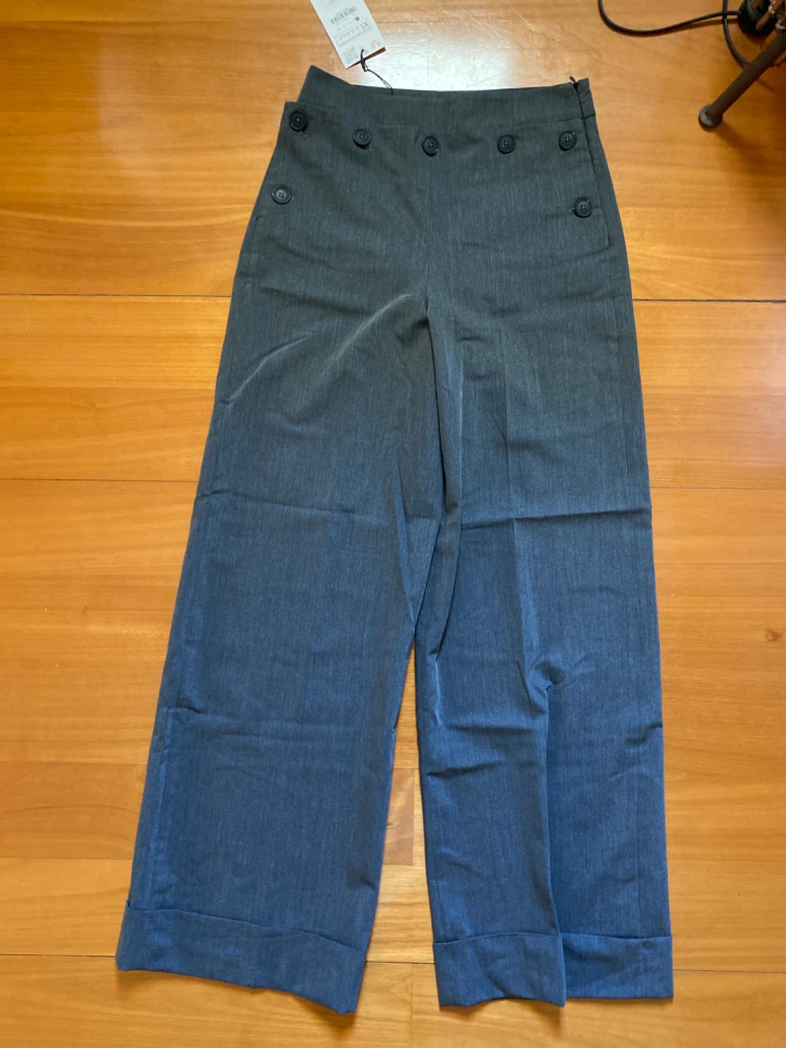 Grey wide-leg trousers with black buttons at the waist, on a light wooden floor. Label visible at the top.