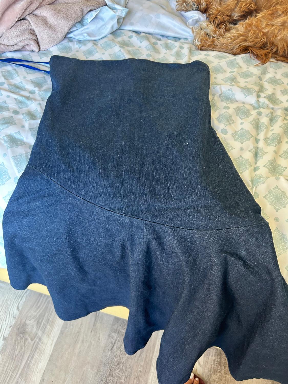 Dark blue denim dress with an asymmetrical hem on a bed. In the background, patterned sheets and a resting dog. Casual setting.