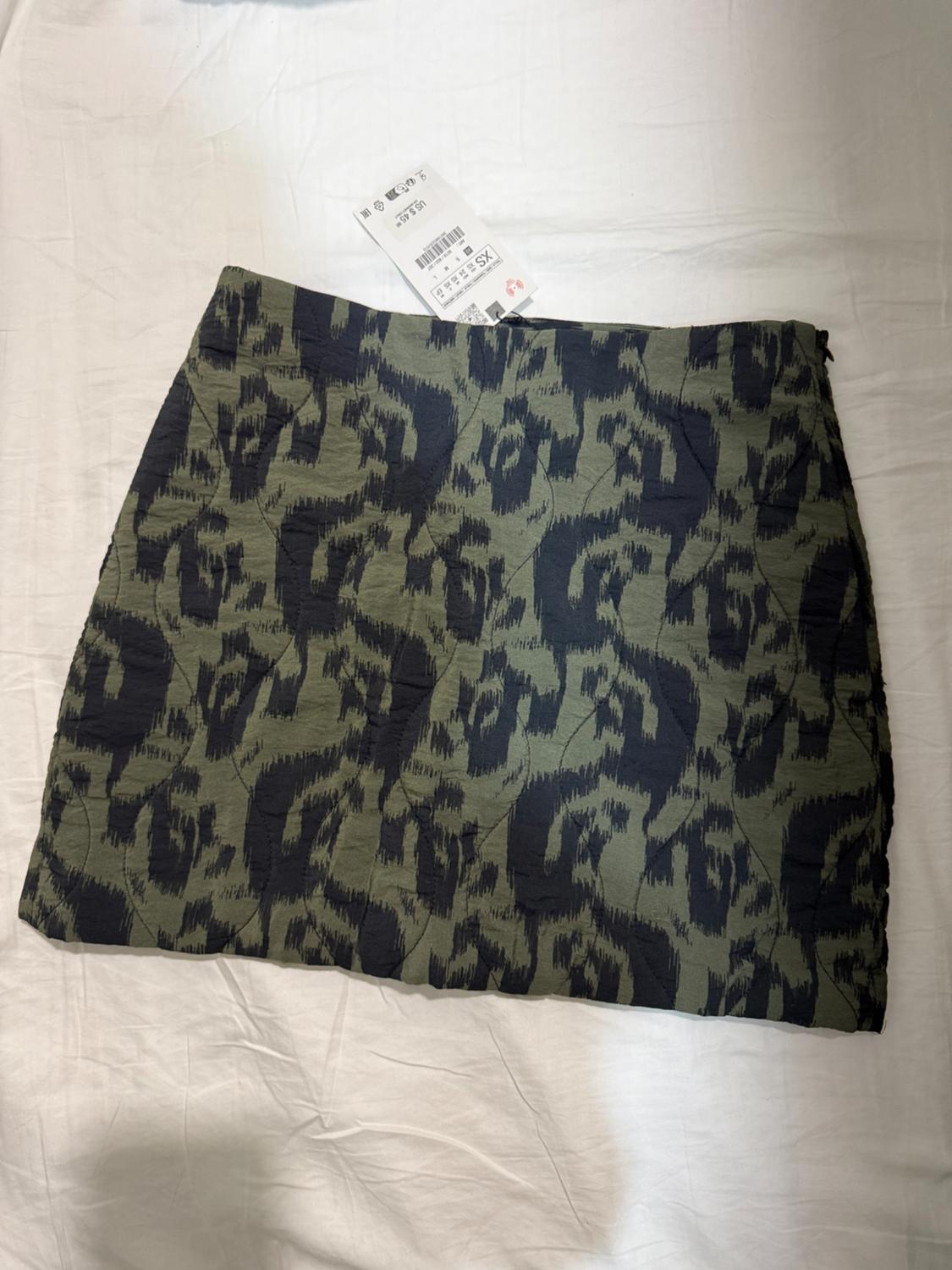 Short skirt with an abstract print in green and black tones on a white background. Visible label with product details.