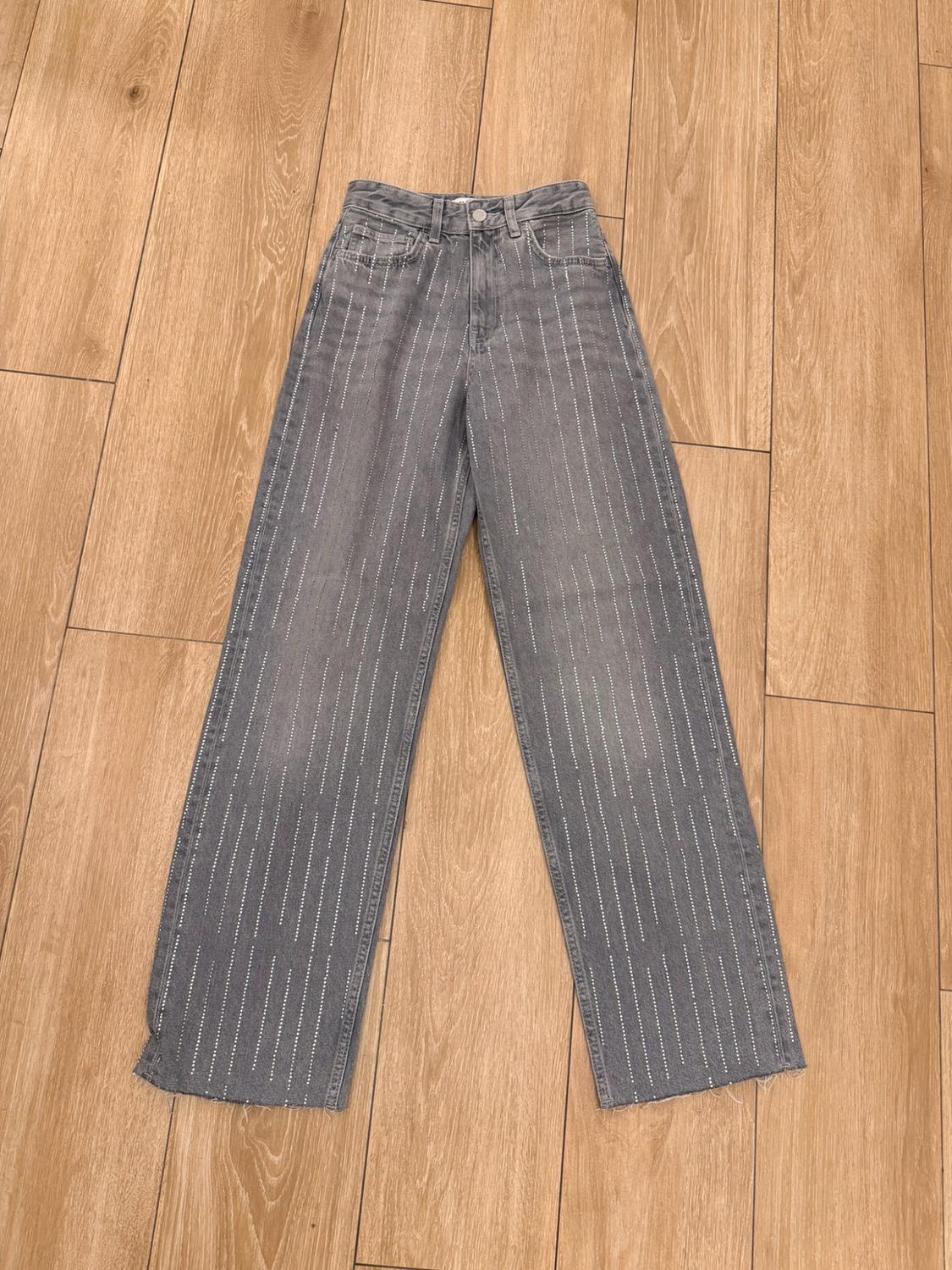 Grey denim trousers with vertical lines of sparkling rhinestones, displayed on a light wooden floor.