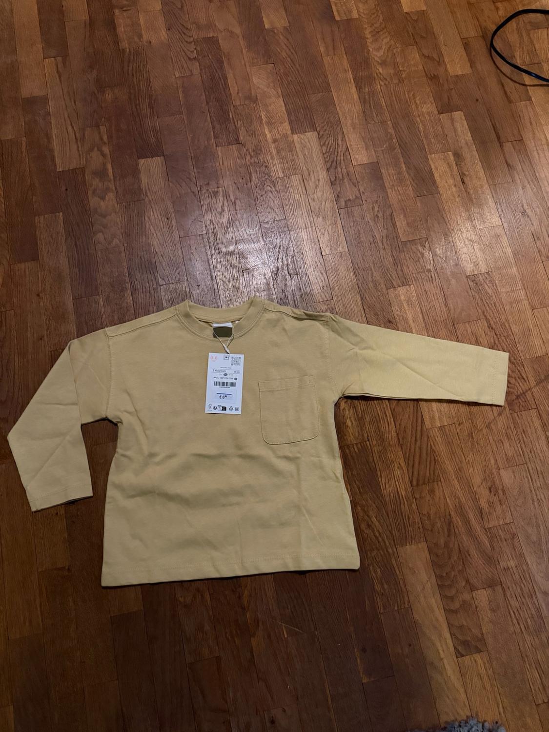 Yellow long-sleeved T-shirt with a front pocket and a label bearing the text “€ 6”, laid out on a wooden floor.