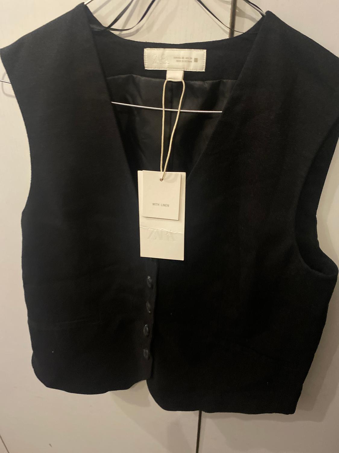 Black waistcoat hanging on a hanger with a white label that says "WITH LINEN". White background.