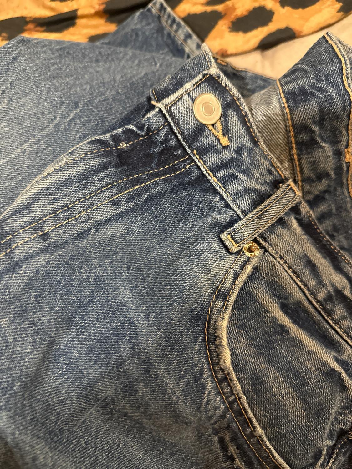 Close-up of blue jeans showing the waistband, metal button, stitching and pocket. In the background, fabric with an animal print can be seen.