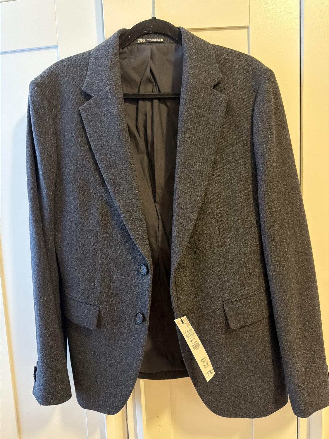 Dark grey blazer on a black hanger against a white door. Interior label with the text ZARA. Casual design with two buttons and pockets.