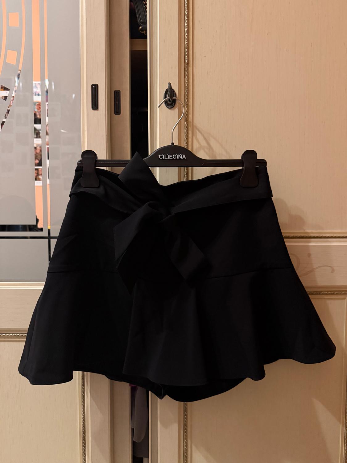 Black skirt with a large bow hanging on a black hanger with the brand "CILIEGINA", against a beige wardrobe door.