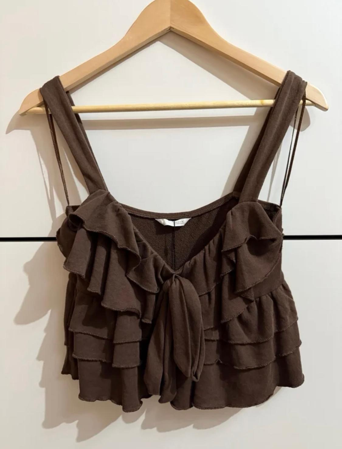 Brown top with ruffles and wide straps, hanging on a wooden hanger against a white background.