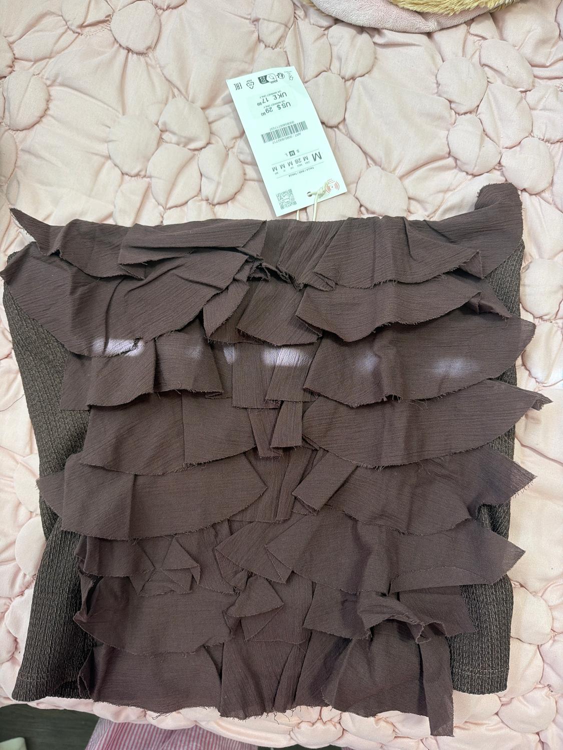 Brown throw pillow with ruffles on pink quilted bedspread. Visible tag with size and price details in dollars and pounds.